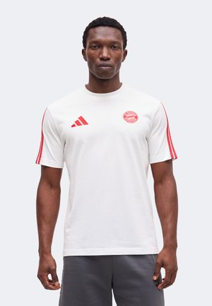 Man draagt een wit sportshirt met korte mouwen, rode Adidas-strepen op de mouwen en het FC Bayern München-logo op de borst, staand tegen een lichte achtergrond.