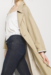 Trench coat de lino beige con un diseño estructurado, superpuesto sobre una blusa blanca y jeans de mezclilla oscuros. El abrigo cuenta con un cuello clásico y un corte holgado.
