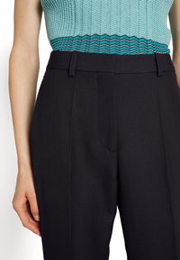 Czarne, dopasowane spodnie z gładkiego materiału, z płaskim przodem i eleganckim pasem. Pasta dodaje subtelnej tekstury i kształtu.