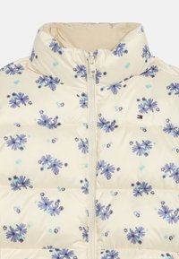 Polstret jakke i krem med lilla blomstermønster. Har høy krage, frontlomme med glidelås og lett, blankt stoff.