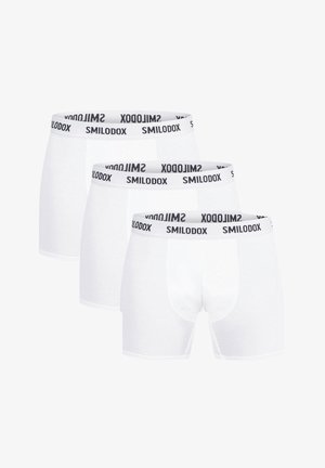 Białe bokserki z czarnym elastycznym pasem, z logo "SMILODOX". Model stoi boso, podkreślając design spodenek.