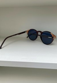 Runda solglasögon med mörkblå linser, bruna och transparenta bågar samt svarta skalmar. Har silverdetaljer och varumärkesdetalj på skalmen.