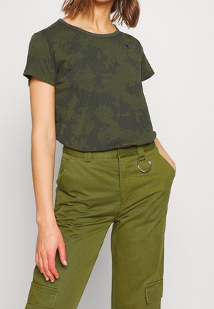 Vrouw draagt een donkergroen T-shirt met ruitpatroon, ingestopt in olijfgroene broek met een grote metalen ring als accessoire aan de riemlus.