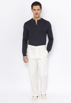 Marineblauwe longsleeve henleyshirt, gecombineerd met witte broek en beige instappers. Eenvoudig ontwerp, zachte textuur, getailleerde pasvorm en knoopaccenten.