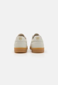Vita lädersneakers med vadderad krage, tan gummisula och guldlogodetalj på hälen. Slät yta och rundad form.