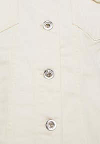 Ljus beige denimjacka med silverfärgade tryckknappar, som har dubbla bröstfickor och sydda sömmar. Mjuk, texturerad tygdetalj synlig.