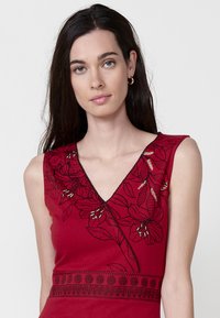 Rotes ärmelloses Kleid mit V-Ausschnitt, floralen Stickereien und strukturierten Mustern entlang des Saums und der Schultern.