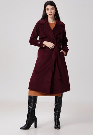 Un manteau trench en laine bordeaux avec une taille ceinturée, de larges revers et des accents en boutons, porté sur une robe côtelée marron et des bottes noires à hauteur de genoux.