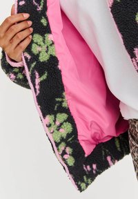 Čierna fleecová bunda s ružovými a zelenými kvetinovými vzormi. Podšitá mäkkým ružovým materiálom. Má kontrastný zips a textúrový povrch.