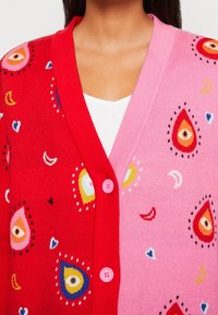 Cardigan rouge et rose avec des motifs d'œil et de cœur, doté de boutons ronds et d'une texture douce. Ornements colorés partout.