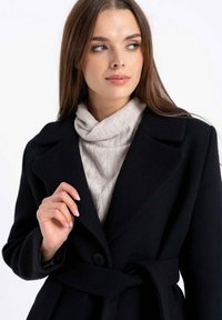 Czarny wełniany płaszcz z paskiem i szerokimi klapami, nałożony na beżowy, teksturowany golf. Modelka ma długie włosy, a tło jest neutralne.