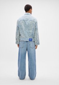 Man wearing textured light blue jacket and wide-leg blue jeans, standing facing away against plain white background.