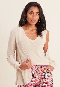 Maglione lavorato a maglia beige con scollatura a V profonda e drappeggio laterale, indossato sopra pantaloni decorati colorati con vortici nei toni del rosso e del blu.