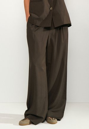 Pantalon marron à larges jambes avec rayures fines porté avec un blazer assorti et des chaussures beiges décontractées sur une personne debout les mains dans les poches.