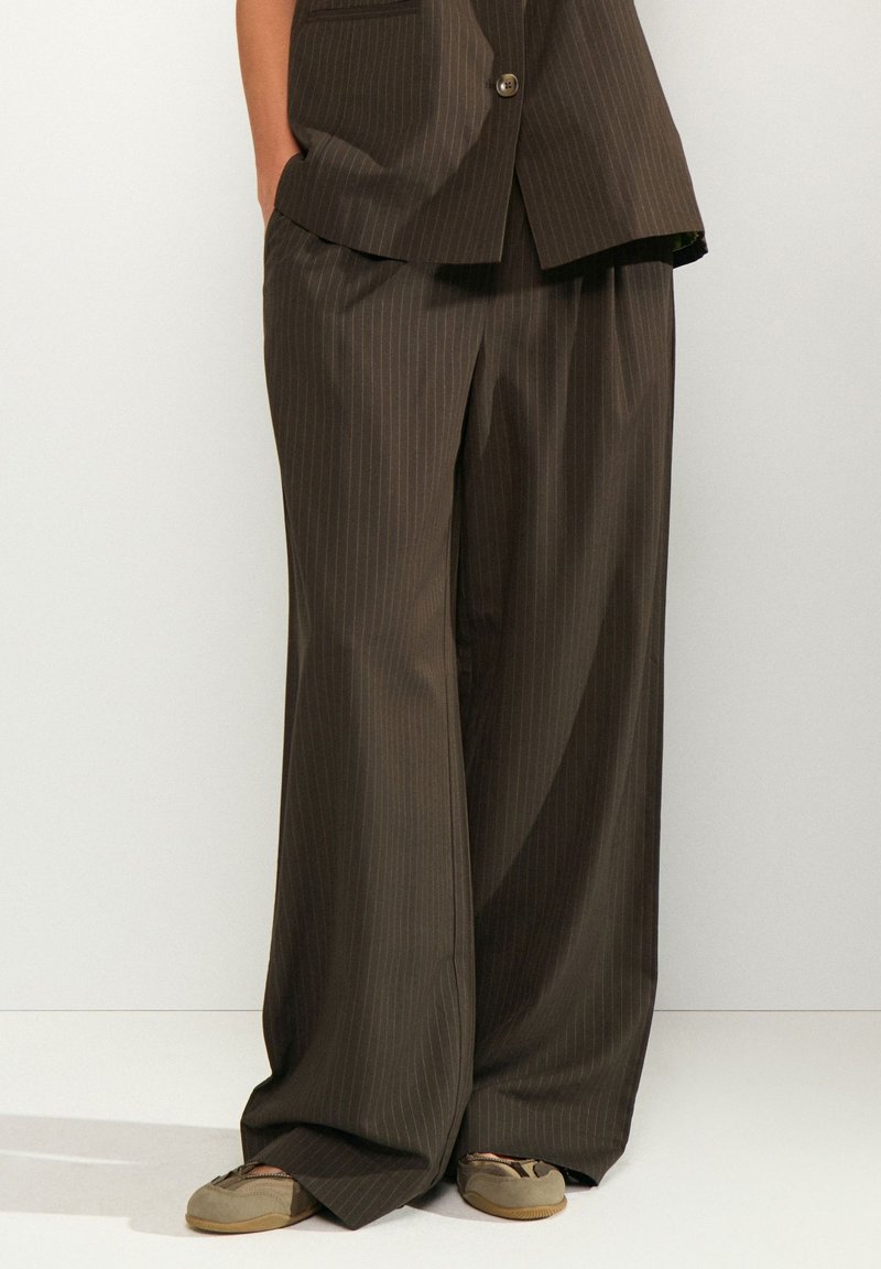 Pantalon marron à larges jambes avec rayures fines porté avec un blazer assorti et des chaussures beiges décontractées sur une personne debout les mains dans les poches.