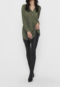 Tunique vert olive à col en V avec trois boutons, associée à des leggings en similicuir noir, présentant une coupe longue et ample.