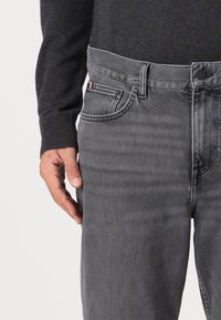 Hombre vestido con un suéter negro y jeans de mezclilla grises, con una pequeña etiqueta de tela roja, blanca y azul cerca del bolsillo delantero, con la mano relajada a un lado.