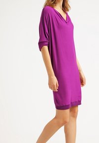 Lila klänning med V-ringning, sänkt axeldesign och korta ärmar. Tillverkad av mjukt tyg, har den en rak passform och kortare fåll.