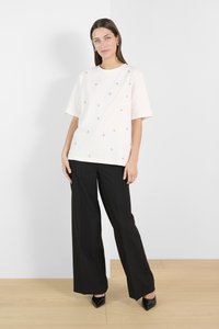 Tricou oversized crem cu nasturi metalici decorativi, asociat cu pantaloni largi negri și pantofi cu vârfuri negre, în fața unui fundal simplu.
