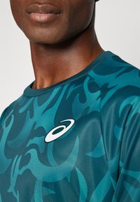 Sinine sportlik särk abstraktse mustriga, ümar kaelus ja lühikesed varrukad. Rinnal on valge logo aktsent. Sile, kerge kangas.