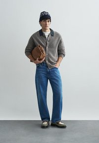 Γκρίζα πλεκτή κάρντιγκαν πάνω από ένα άσπρο πουκάμισο, μπλε τζιν και καφέ παπούτσια. Κρατώντας μια καφέ τσάντα και φοράει ένα μπλε καπέλο με σχέδιο.