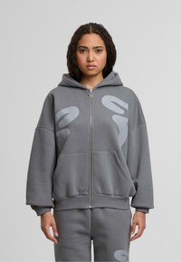 Sudadera gris con cremallera, de corte relajado, con grandes bolsillos frontales, capucha y patrones abstractos en gris claro en la parte delantera. Hecha de una tela suave.