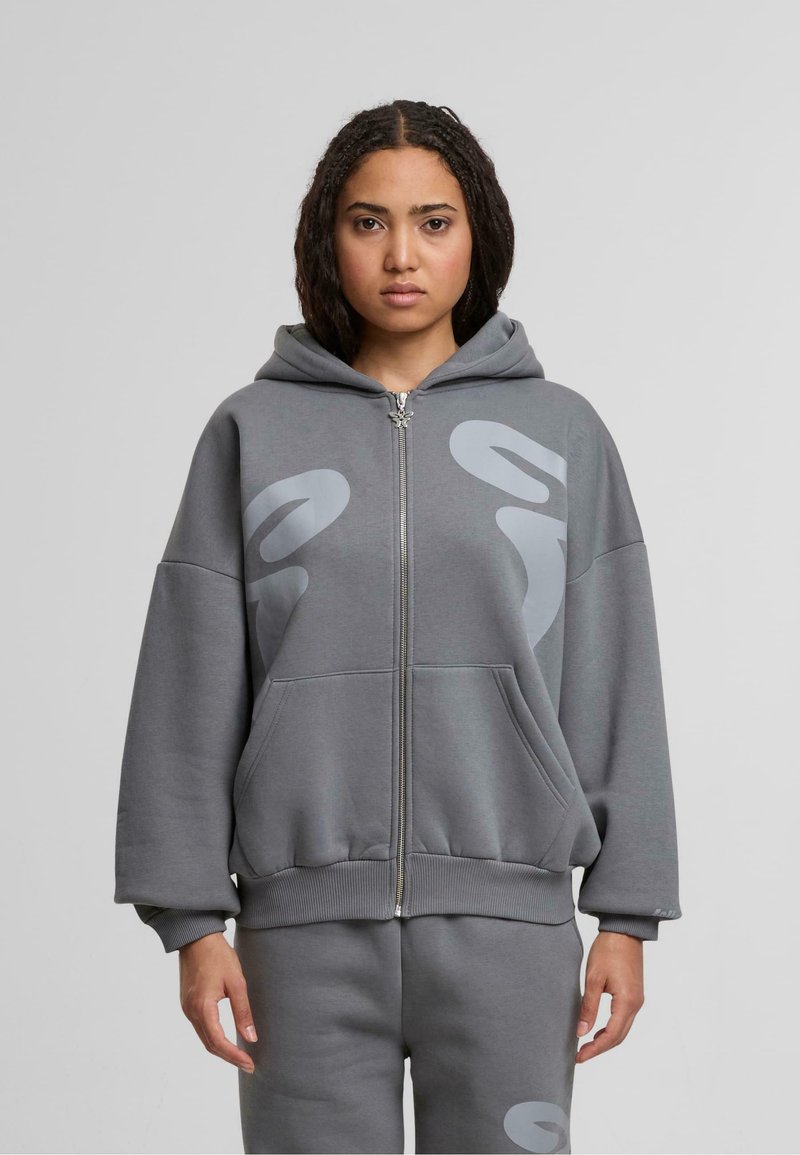 Sudadera gris con cremallera, de corte relajado, con grandes bolsillos frontales, capucha y patrones abstractos en gris claro en la parte delantera. Hecha de una tela suave.