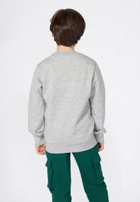 Bambino con capelli castani corti che indossa un felpa grigia chiara e pantaloni cargo verdi, in piedi rivolto di spalle contro uno sfondo bianco.