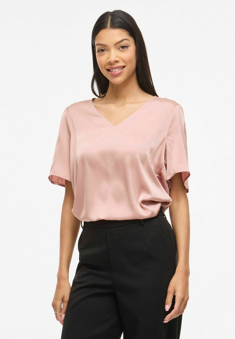 Femme aux longs cheveux noirs portant un chemisier en satin rose clair et un pantalon noir taille haute, debout et souriante sur un fond blanc.