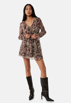 Vrouw draagt een bruine gestippelde lange mouw met ruches mini-jurk en zwarte kniehoge laarzen met hak, staand tegen een effen achtergrond.