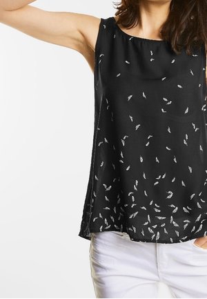Femme portant un haut noir sans manches avec de petits motifs de plumes blanches et un pantalon blanc, bras levé.
