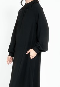 Crna haljina dugih rukava s visokim izrezom, nabranim detaljima i bočnim džepovima. Izrađena od glatkog, teksturiranog materijala.