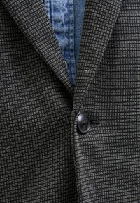 Schwarze und graue gemusterte Blazerjacke mit Revers und einer einzelnen Knopfverschluss, über einem blauen Denim-Hemd getragen.