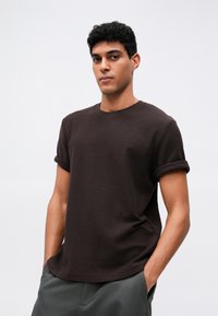 Mladý muž s kučeravými vlasmi, ktorý má na sebe tmavohnedé tričko s vyhrnutými rukávmi a šedé nohavice, stojí so založenými rukami v vreckách na pozadí s rovnomerným vzhľadom.