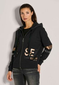 Schwarze Zip-Hoodie mit metallischen Akzenten, versehen mit dem goldenen Druck "SEE YOU SOON". Hergestellt aus weichem Stoff; beinhaltet eine Kapuze und gemusterte Ärmel.