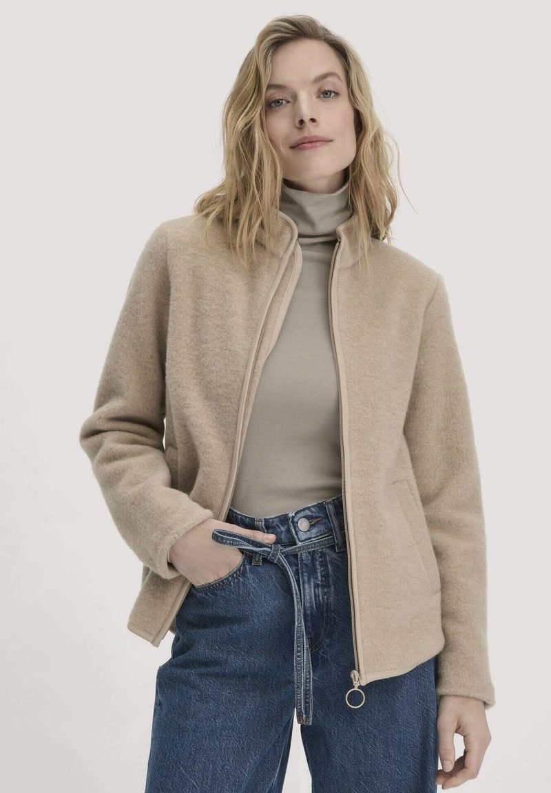 Beige Wollmischjacke mit Reißverschluss, hohem Kragen und Seitentaschen, kombiniert mit einem figurbetonten beigen Rollkragenpullover und blauen hochgeschnittenen Jeans.