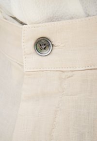 Beige fabric with a textured surface. Features a round, dark button with four holes and contrasting stitching along the seam.