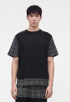 Jeune homme portant un t-shirt noir aux manches et à l’ourlet à motifs, debout devant un fond clair uni, faisant face.