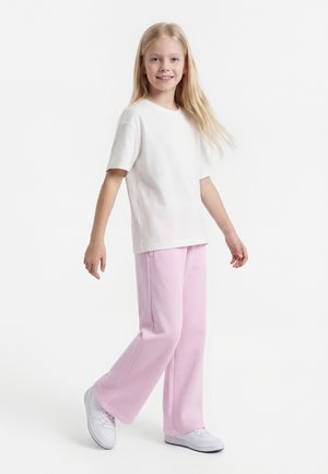 Jeune fille aux longs cheveux blonds portant un t-shirt blanc, un pantalon rose et des baskets blanches, marchant avec un léger sourire.