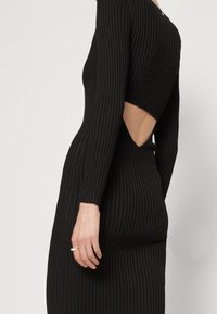 Abito nero a coste con maniche lunghe, caratterizzato da un'apertura sul retro. Texture liscia, forma aderente e design minimalista.