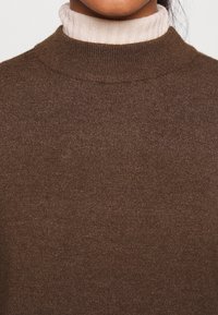 Maglione a collo alto in maglia marrone con una texture liscia, caratterizzato da un colletto alto e un collo alto di crema sovrapposto sotto.