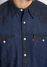 Camisa de mezclilla azul oscuro con un diseño de yugo en contraste, botones a presión y dos bolsillos frontales con una etiqueta roja de Levi's en el bolsillo izquierdo.