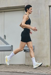 Des chaussures de sport blanches et jaunes avec des semelles noires, assorties avec des chaussettes blanches et un crop top noir avec un short, portées par une personne en train de courir.