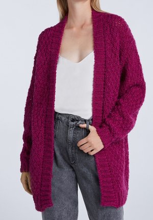 Frau trägt eine strukturierte, tiefmagentafarbene Strickjacke über einem weißen Top und hoch taillierte, ausgewaschene schwarze Jeans, eine Hand in der Gürtelschlaufe eingesteckt.