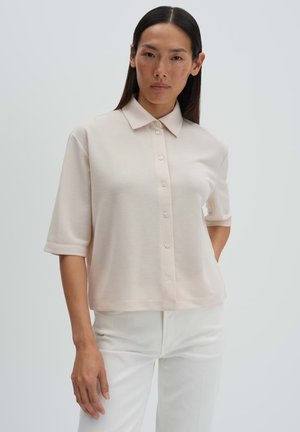 Frau mit langen dunklen Haaren, die ein hellbeiges kurzärmeliges Hemd mit Knöpfen und weiße Hosen trägt, steht vor einem schlichten hellen Hintergrund.