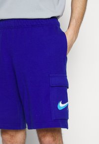 Homme portant un short de sport bleu vif avec une poche latérale et le logo Nike, la main reposant à l'intérieur de la poche, sur un fond blanc uni.