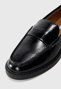 Sorte patentlær loafers med en glat, blank overflade, der har et syet design og subtile detaljer langs siderne.