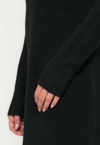 Černé pletené šaty s dlouhými rukávy a žebrovanými manžetami. Měkká textura s mírně volným střihem, zdůrazňující minimalistický design.