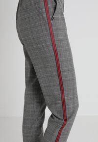 Grå rutiga byxor med slim fit, prydda med en röd och grön sidrand. Tillverkade av en tyg med slät textur. Främre fickor synliga.