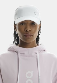 Witte pet met geperforeerde zijkanten en een gebogen rand, gedragen met een lichtroze hoodie met een opvallend logo en trekkoorden.
